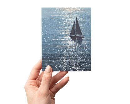 Hand holding a greeting card with an illustration of a sailboat in silhouette drifting through sparkling blue water, with sunlight reflecting in shimmering streaks and dots. The card features a dreamy, ocean-inspired palette with a painterly texture.