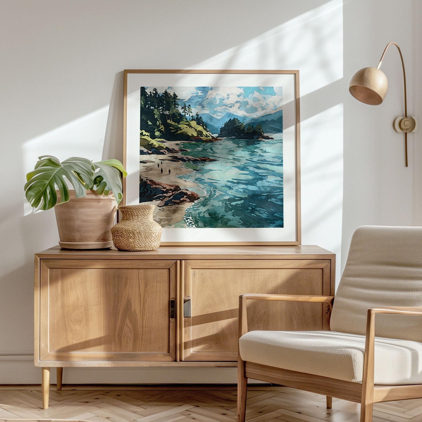 Framed coastal landscape art print displayed on a wooden sideboard in a bright, modern living space. The artwork shows turquoise ocean waves, sandy shore, rocky cliffs, and evergreen trees beneath a partly cloudy sky. The room features a potted monstera plant, woven vase, beige armchair, and wall-mounted lamp, with sunlight casting soft shadows across the scene.