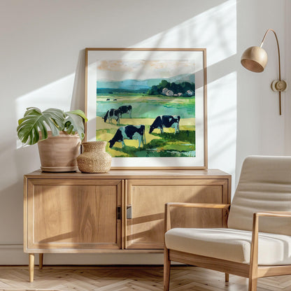 Framed modern impressionist painting of cows grazing in a green meadow, with rolling blue hills and white barns in the distance. The artwork rests on a wooden sideboard in a bright, minimalist living space, styled with a potted plant, woven vase, neutral armchair, and wall-mounted lamp. Sunlight filters through, casting soft shadows for a warm, natural atmosphere.