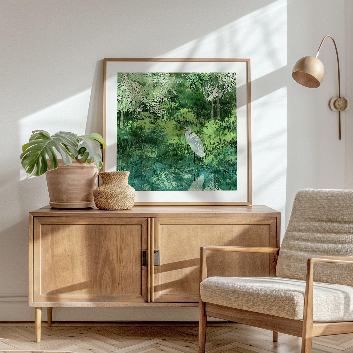 Square-framed art print of a white egret beside a still pond, surrounded by dense greenery and delicate white blossoms. The watercolor-style artwork is styled in a modern, sunlit room with mid-century furniture, a monstera plant in a clay pot, and a woven basket for a natural, serene aesthetic.