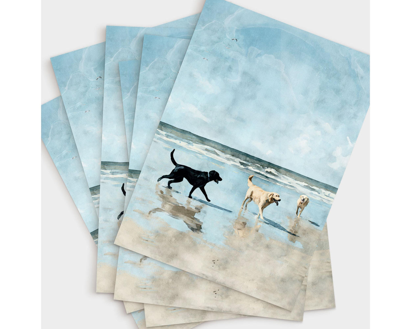 Set of greeting cards featuring a watercolor-style illustration of three dogs—one black and two golden—playing on a sandy beach with gentle waves under a bright blue sky, fanned out in an overlapping arrangement on a light background.