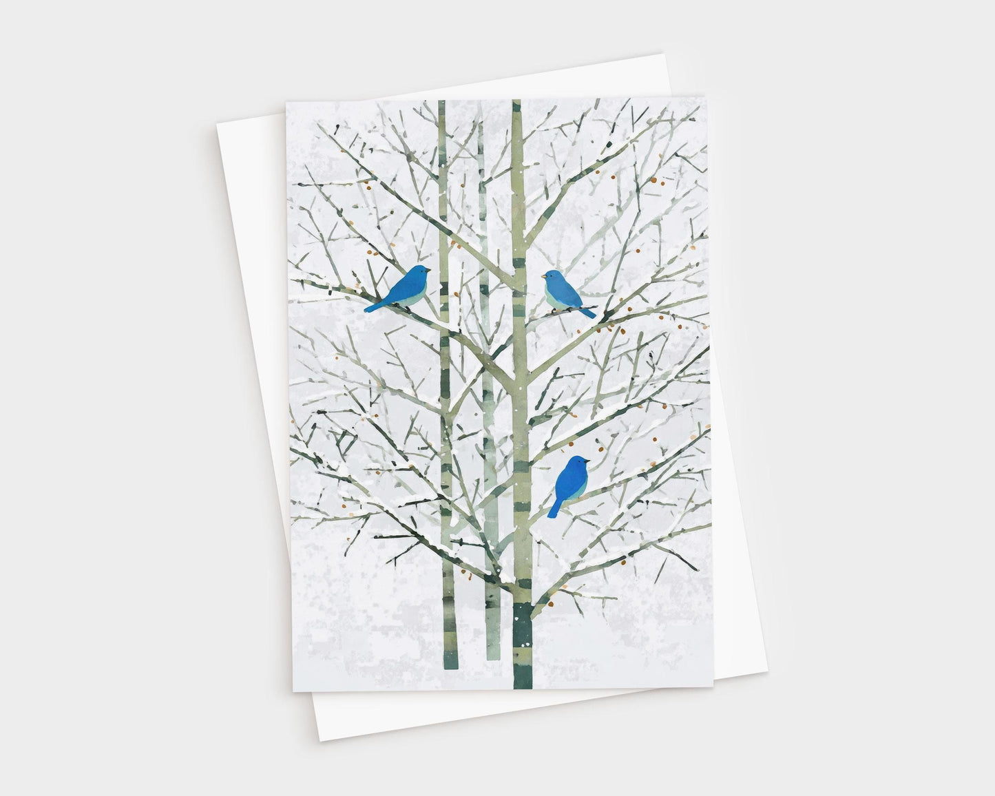 Greeting card featuring a minimalist watercolor-style illustration of three bright bluebirds perched on bare winter branches with pale green trunks and a soft white background. The card is shown with a white envelope, creating a natural, elegant presentation.