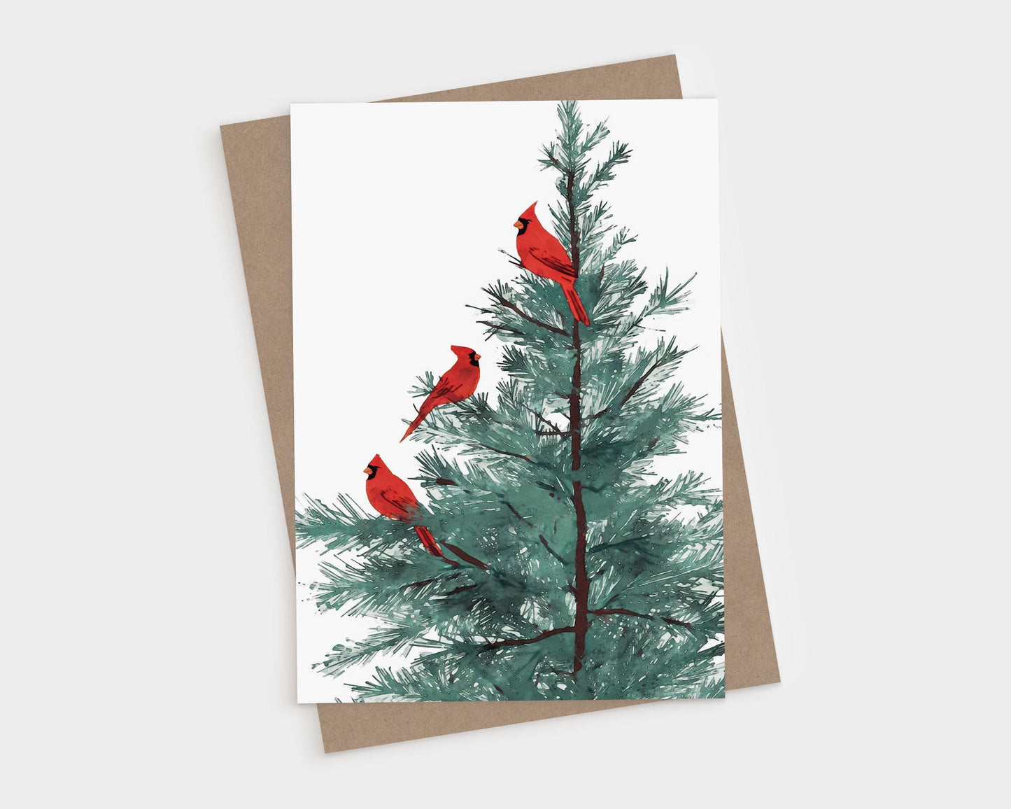 Greeting card featuring an illustration of three bright red cardinals perched on the branches of a green pine tree, set against a clean white background. A kraft brown envelope is placed behind the card.