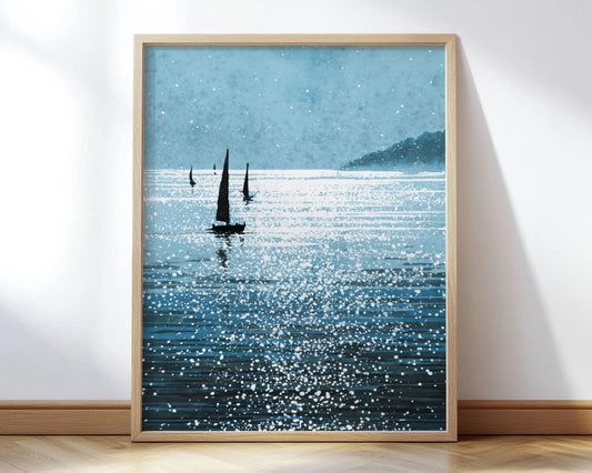 Framed art print featuring a serene seascape with silhouetted sailboats gliding across sparkling blue water. White flecks mimic sunlight reflecting off gentle waves, creating a dreamy, almost abstract texture. A misty headland appears in the background beneath a pale sky. The piece is displayed in a light wood frame, leaning against a white wall on a wood floor.