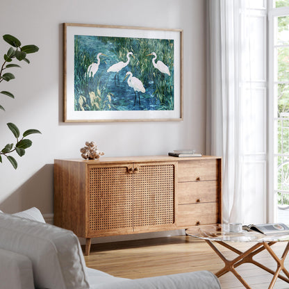 Framed painting of five white egrets in a marsh, with blue water and tall green grasses, hangs above a mid-century style wood sideboard with woven rattan doors. The bright, airy living room features a white sofa, glass coffee table, books, and large windows with sheer curtains, creating a calm, natural atmosphere.