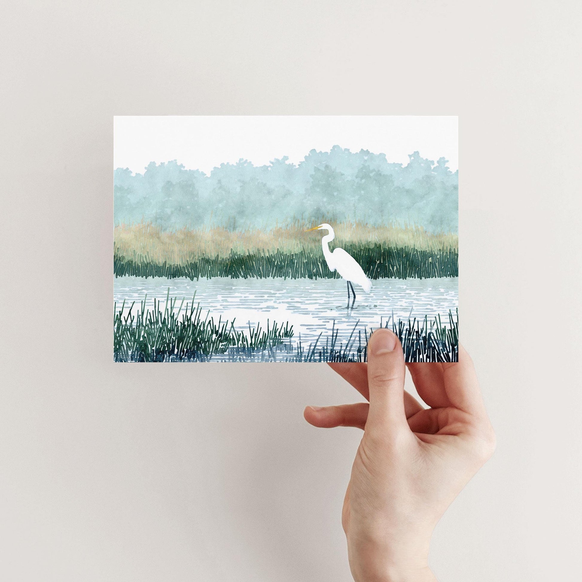Hand holding a greeting card featuring a watercolor-style illustration of a white heron standing in a marsh, surrounded by tall green grasses, calm water, and misty blue-green trees in the background.