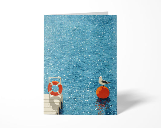 Greeting card standing upright on a white surface, featuring a minimalist watercolor-style illustration of a seagull perched on a bright orange buoy in sparkling blue water. A small white dock with a red life preserver appears in the corner, adding nautical charm to the serene coastal scene.