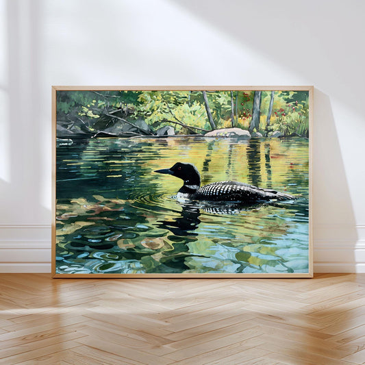Framed art print of a loon gliding across a calm lake surrounded by lush green trees. The water reflects shades of gold, green, and blue, capturing the bird’s distinctive black-and-white plumage and the dappled sunlight filtering through the forest. The print rests against a white wall on a light wood floor, bathed in soft natural light.
