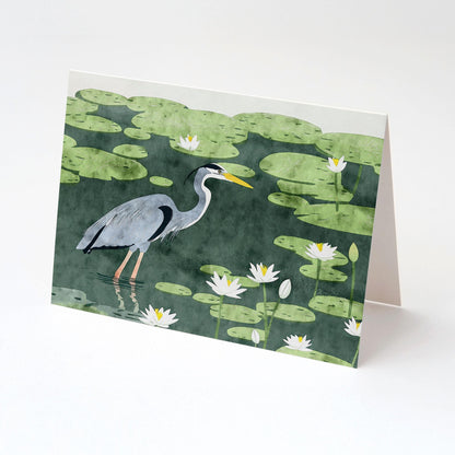 Folded greeting card featuring a painted illustration of a great blue heron standing in a pond among green lily pads and blooming white water lilies. The card is photographed against a clean white background, shown at an angle to display its blank interior.
