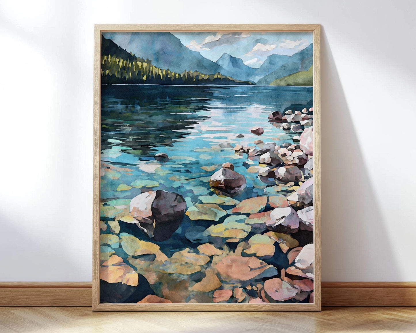 Framed watercolor painting of a serene lake with crystal-clear water revealing colorful rocks beneath the surface, surrounded by mountains and pine trees under a partly cloudy sky.