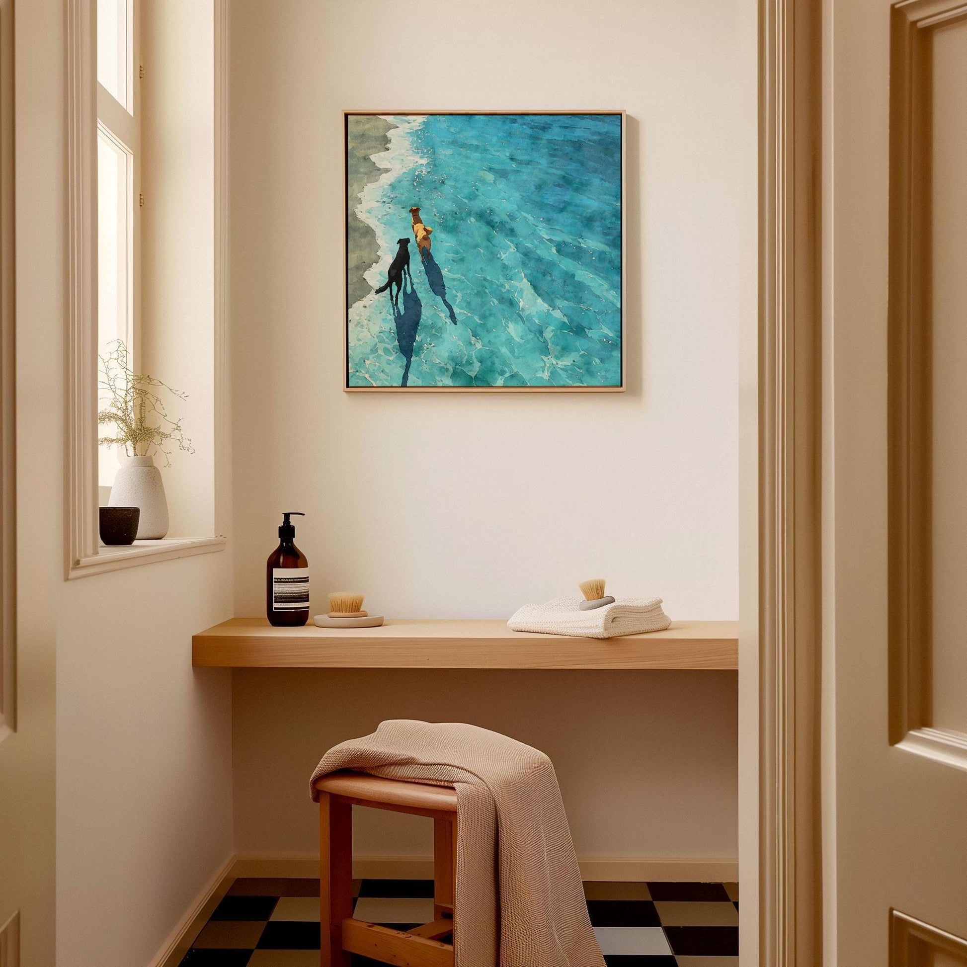Square framed art print of two dogs walking along the shoreline, one golden and one black, their shadows stretching into sunlit turquoise water. Displayed above a minimalist wooden bench in a serene, spa-like room, the watercolor piece adds a calm, coastal charm to the space.