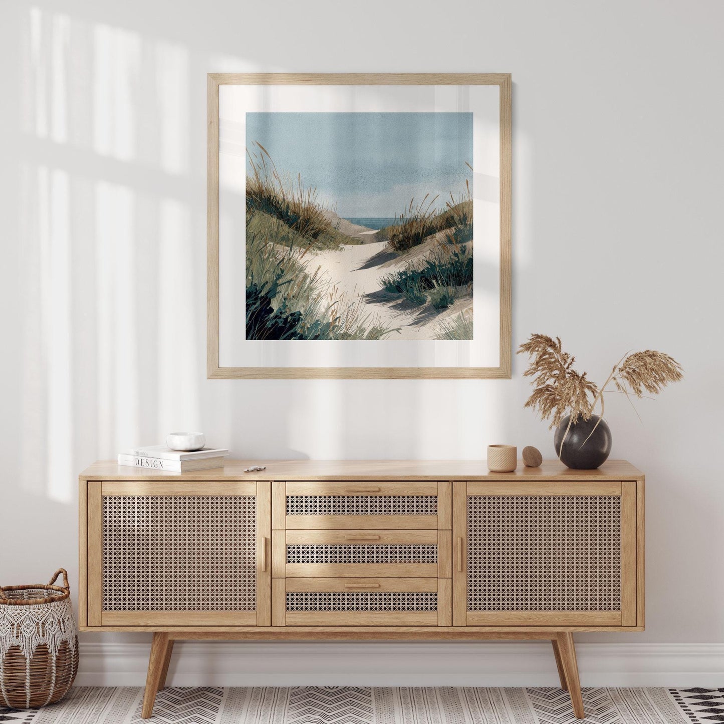 Square framed coastal watercolor print of a sandy dune path with tall grasses leading to the ocean, displayed above a light wood cabinet with woven rattan doors. Decor includes a black vase with dried grasses, small ceramics, and a basket, in a sunlit minimalist room.