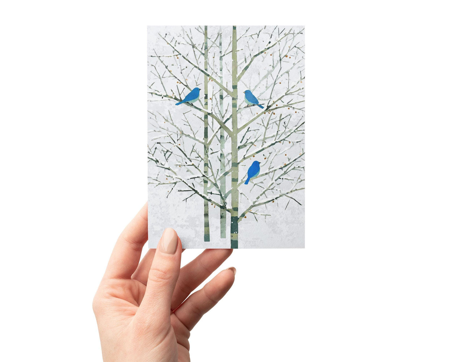 Hand holding a greeting card featuring a minimalist watercolor-style illustration of three vivid bluebirds perched on bare winter tree branches. The delicate green-gray trunks and white background create a serene, modern design perfect for winter or nature-themed occasions.