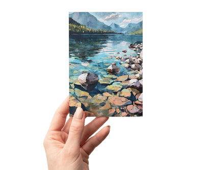 Hand holding a card featuring a watercolor-style illustration of a tranquil mountain lake, with clear blue-green water, smooth colorful rocks in the foreground, forested shoreline, and distant blue mountains under a partly cloudy sky.