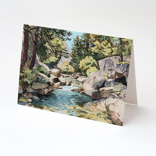 Folded greeting card featuring a watercolor-style painting of a serene mountain stream flowing between large rocks, surrounded by pine trees and sunlit greenery, displayed upright on a white background.