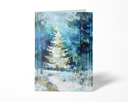 A folded greeting card featuring a watercolor-style painting of a glowing white Christmas tree in a snowy blue-green forest. Soft washes, tall tree silhouettes, and scattered light speckles create a serene winter atmosphere, shown against a clean white background with a subtle shadow.