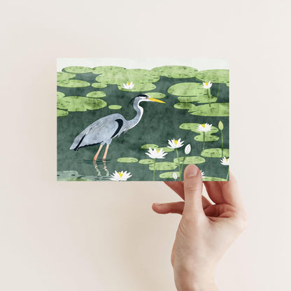 A hand holds a greeting card featuring an illustration of a great blue heron standing in a pond filled with lily pads and blooming white water lilies. The heron is depicted in profile with soft, painterly textures and muted green tones.