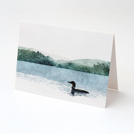 Folded greeting card featuring an illustration of a loon swimming on a calm blue lake with ripples, framed by misty green forested hills beneath a pale sky. The card stands upright against a clean white background, showcasing its front design.