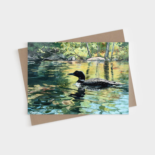 Watercolor-style greeting card featuring a common loon gliding across a calm lake. The bird’s black head and checkered wings reflect in the rippling water, which mirrors the surrounding trees in green and golden tones. Sunlight filters through leaves, creating a serene, nature-inspired scene. The card is displayed with a natural brown kraft envelope on a white background.