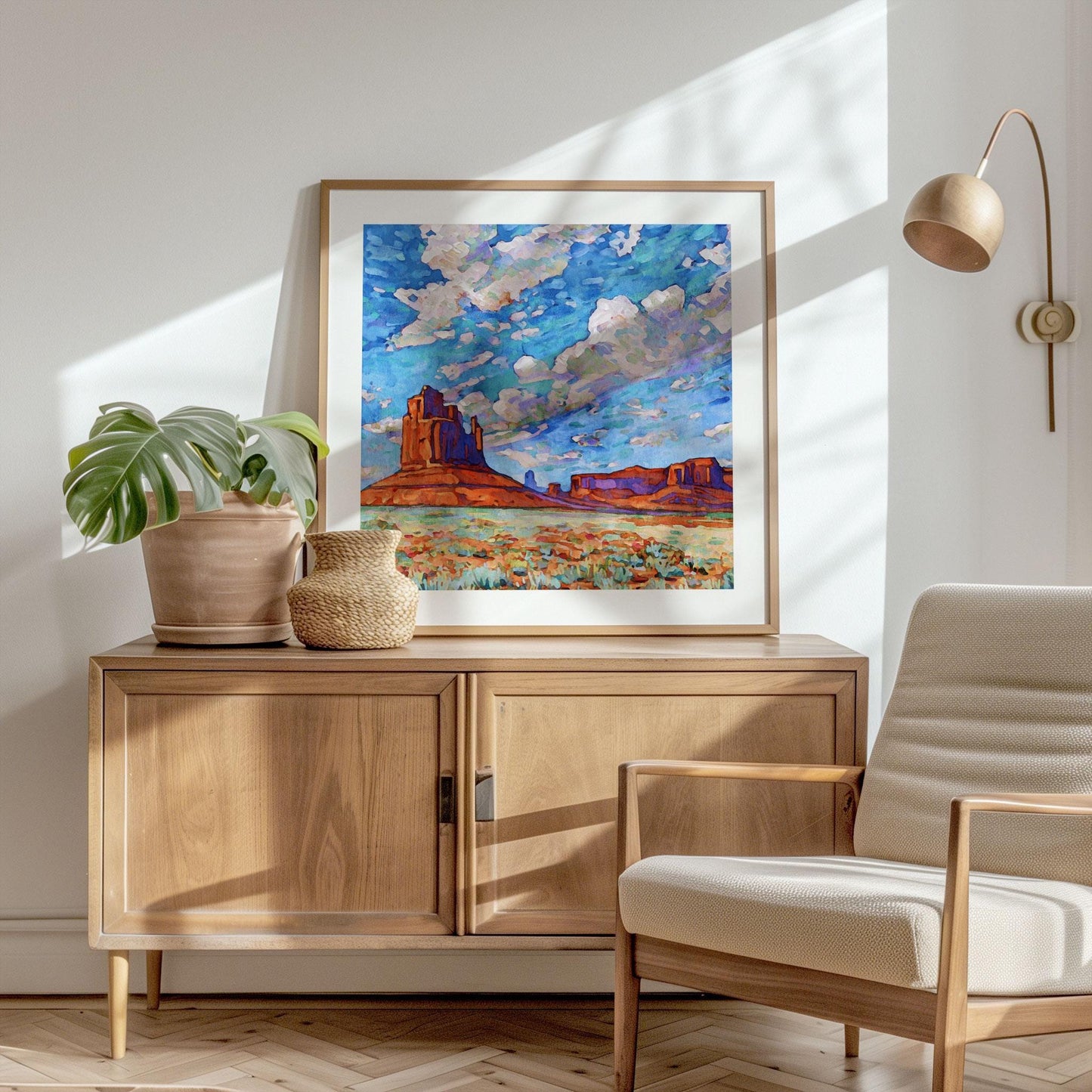 Framed Monument Valley desert painting featuring vivid red rock buttes, green desert plants, and a dramatic blue sky with swirling clouds, displayed on a wooden cabinet beside potted plants and a woven basket in a sunlit modern room.