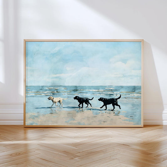 Watercolor-style painting of three Labrador dogs, one yellow and two black, walking along a sandy beach with gentle waves and a bright blue sky in the background, displayed in a light wooden frame leaning against a white wall on a hardwood floor.