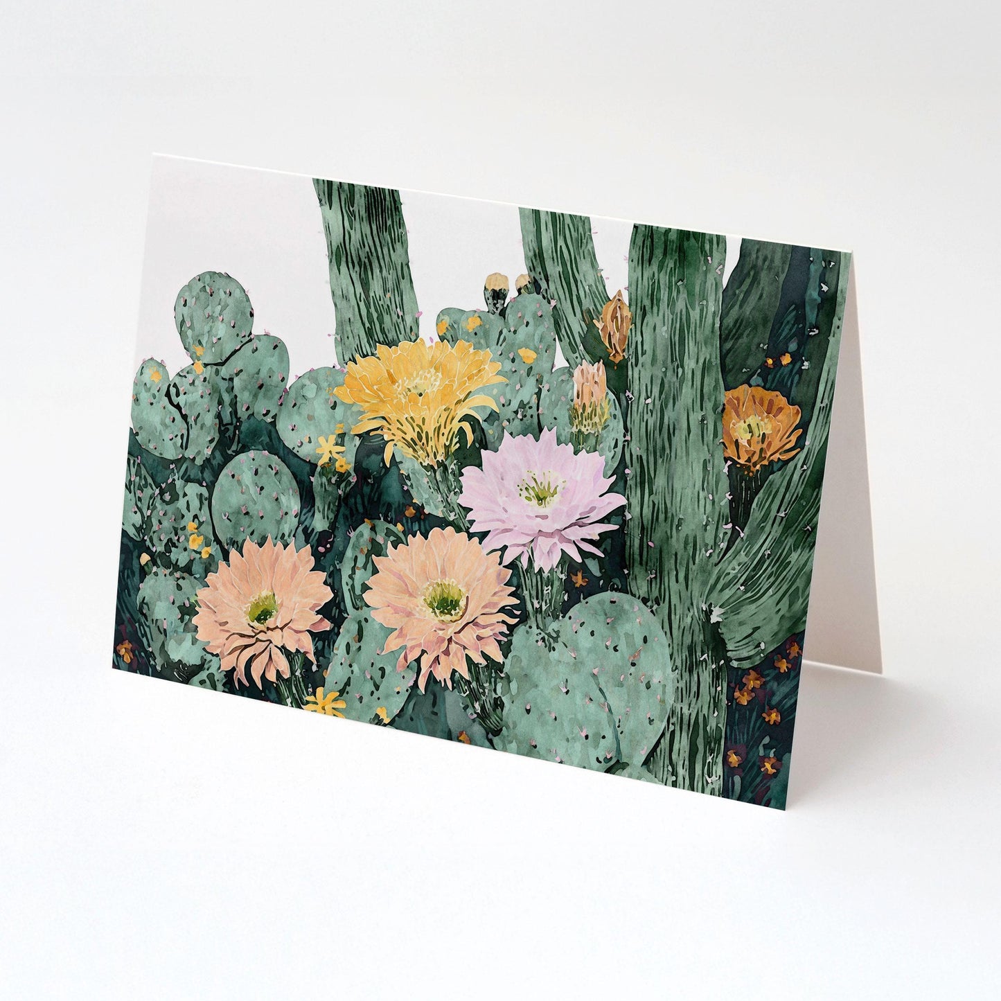 Folded greeting card with watercolor cactus illustration, showing clusters of green pads and tall stems with blooming flowers in peach, pink, yellow, and orange tones, displayed on a clean white background.