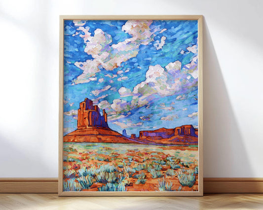 Framed desert landscape painting featuring Monument Valley’s iconic red rock buttes under a dramatic sky filled with swirling blue, purple, and white clouds, with green desert plants in the foreground.