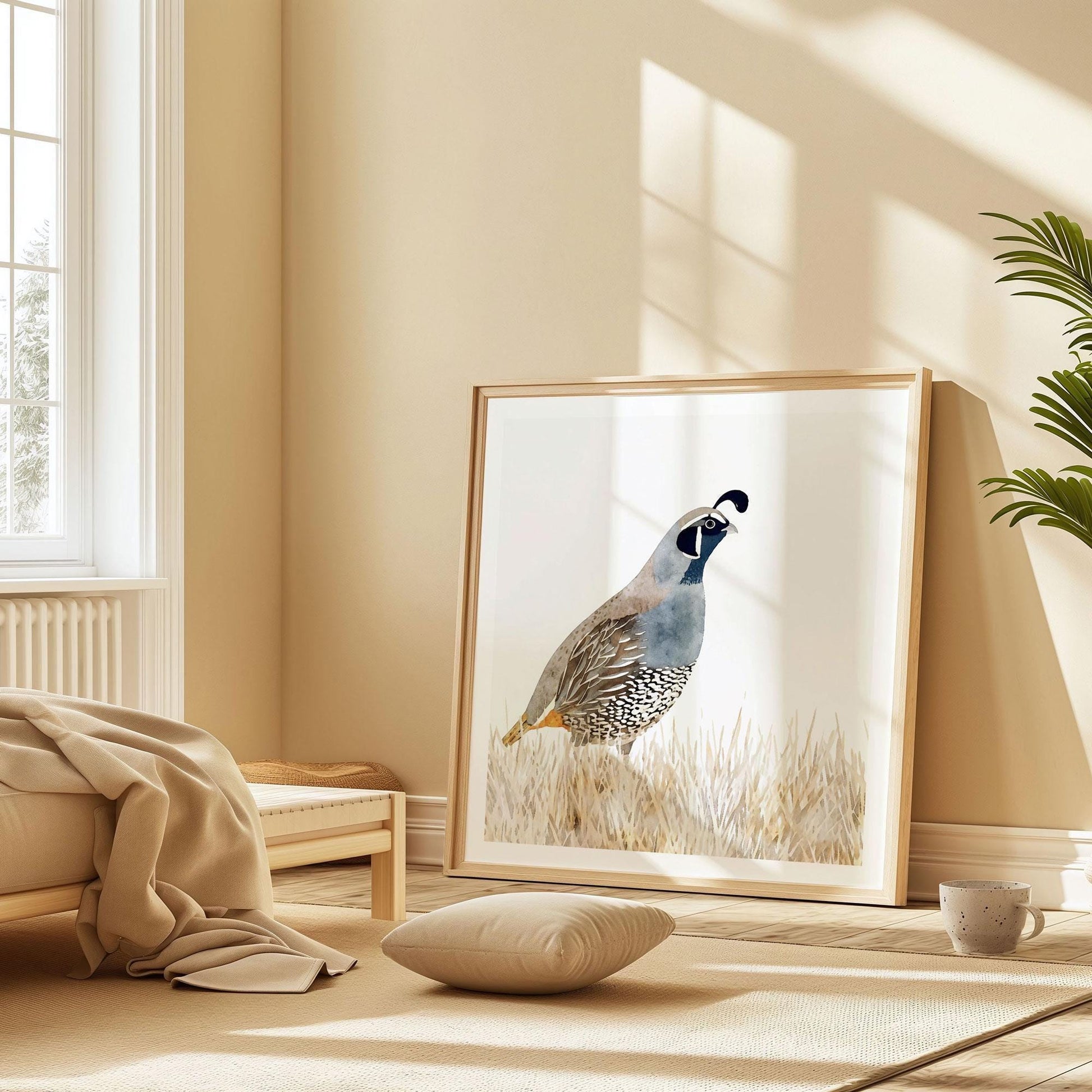 Framed watercolor painting of a California quail rests against a beige wall in a sunlit minimalist bedroom. The artwork sits on the floor beside a low wooden bed with a tan blanket, a cushion, and a ceramic mug, with warm natural light streaming through tall windows.