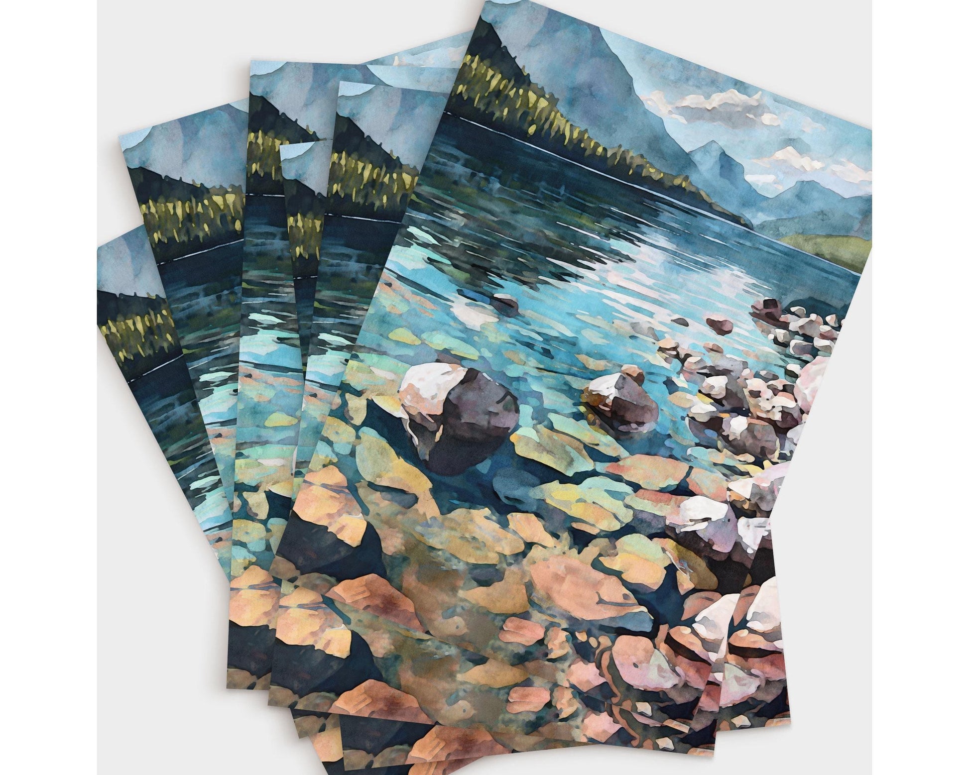 Set of several greeting cards featuring a watercolor-style illustration of a mountain lake with clear water, colorful rocks, a rocky shoreline, forested hills, and distant mountains under a partly cloudy sky, fanned out to show the repeated scenic design.