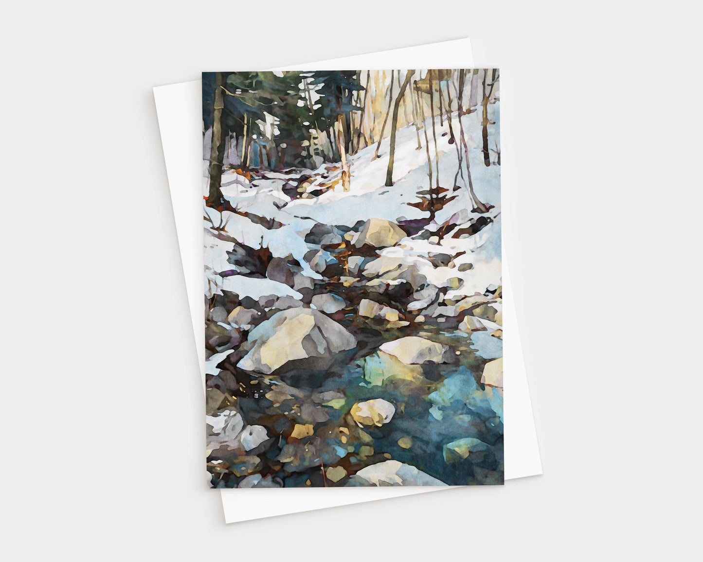 Watercolor-style greeting card featuring a rocky creek running through a snowy forest. Sunlight filters through bare trees, reflecting on the icy water in shades of blue and green. The card is displayed with a white envelope.