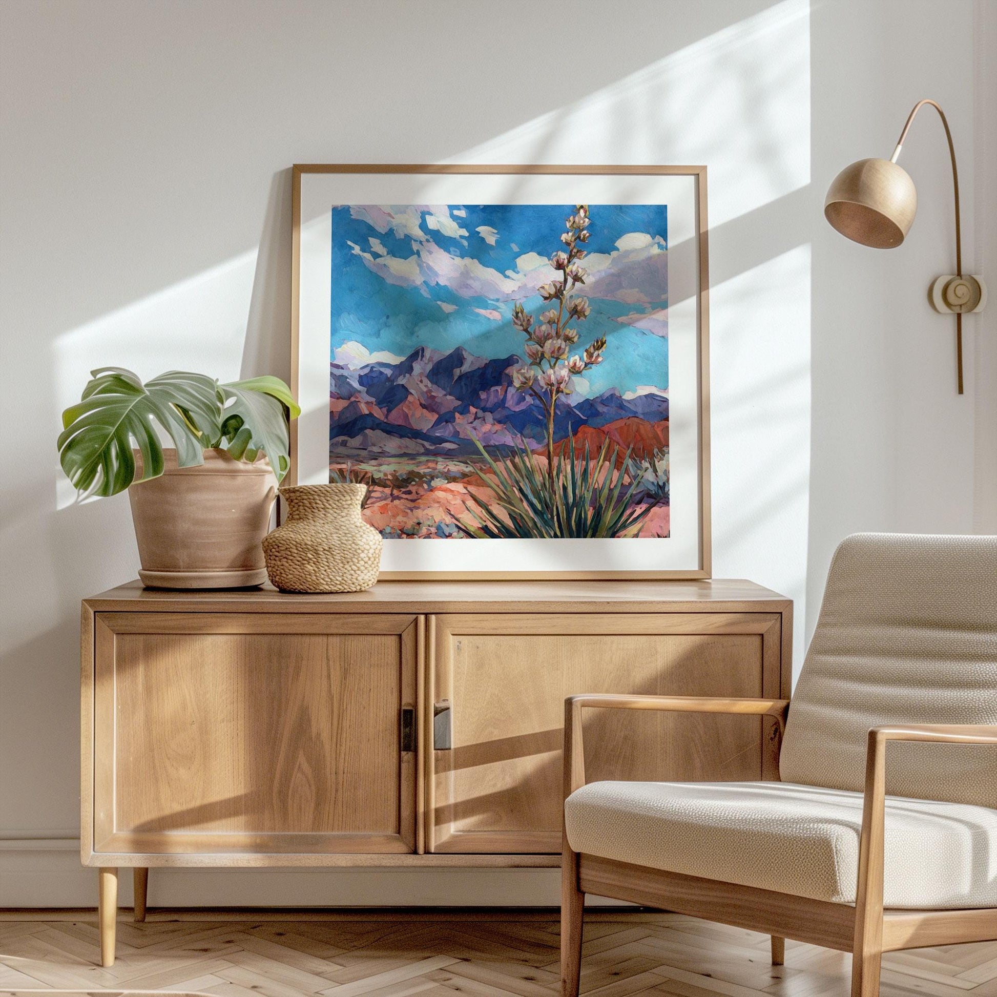 Framed desert landscape painting featuring a tall yucca plant in bloom before purple mountains and a bright blue sky, displayed on a wooden sideboard in a sunlit modern living room. A potted monstera, woven vase, and neutral armchair complete the minimalist, natural decor, with soft light and shadows creating a warm, inviting atmosphere.