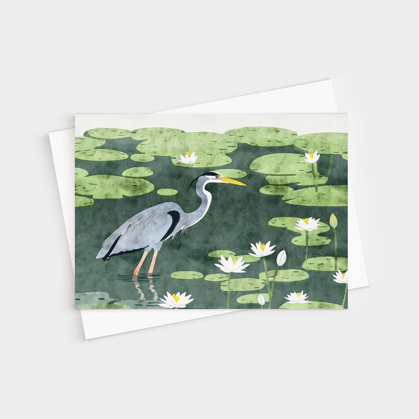 Greeting card featuring an illustration of a great blue heron wading among lily pads and white water lilies in a dark green pond. The heron is shown in profile with detailed feathers and a bright yellow beak. A white envelope is partially visible beneath the card.