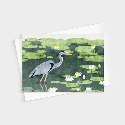 Greeting card featuring an illustration of a great blue heron wading among lily pads and white water lilies in a dark green pond. The heron is shown in profile with detailed feathers and a bright yellow beak. A white envelope is partially visible beneath the card.