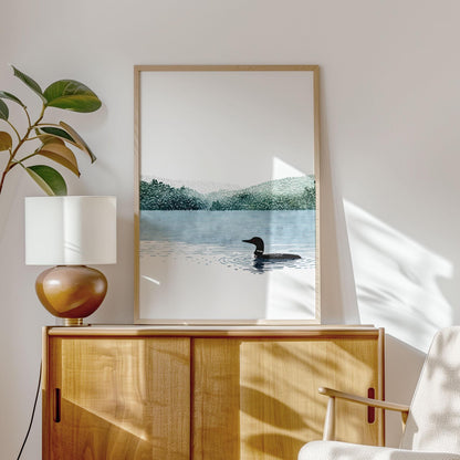 Framed minimalist watercolor art print displayed on a light wood sideboard in a bright, modern interior. The artwork shows a solitary loon swimming on a calm blue lake with soft green forested hills in the distance and a wide white sky. Sunlight casts gentle shadows across the wall, with a potted plant and table lamp beside the frame, creating a serene, nature-inspired home setting.