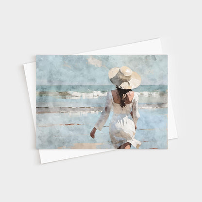 Greeting card featuring a watercolor-style painting of a woman in a flowing white dress and wide-brimmed hat walking toward gentle ocean waves under a soft blue sky.