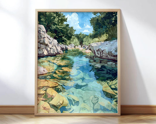 Framed watercolor-style landscape painting of a clear rocky river with turquoise and green water, reflecting sunlight and sky. Large rocks line the shore, with lush green trees in the background under a bright blue sky with clouds. The artwork leans against a white wall on a wooden floor.