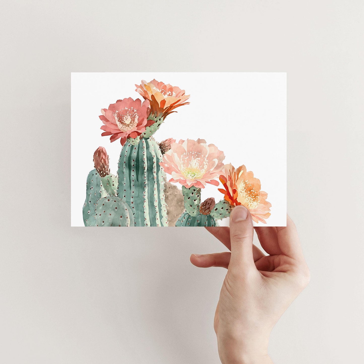 Hand holding a greeting card with a watercolor illustration of green cacti topped with large blooming flowers in shades of pink, peach, and orange against a white background.