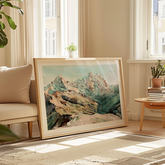 Large framed watercolor mountain painting with soft green, blue, and brown tones, leaning against a wall in a bright living room. The artwork rests on a patterned rug beside a beige sofa and round wooden table with books and plants, sunlight streaming through tall windows.
