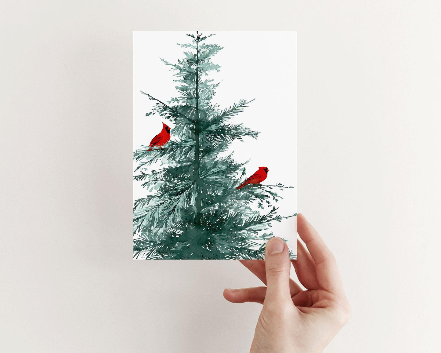 Hand holding a greeting card featuring a watercolor illustration of a pine tree with two vivid red cardinals perched on its branches, displayed against a plain light background.