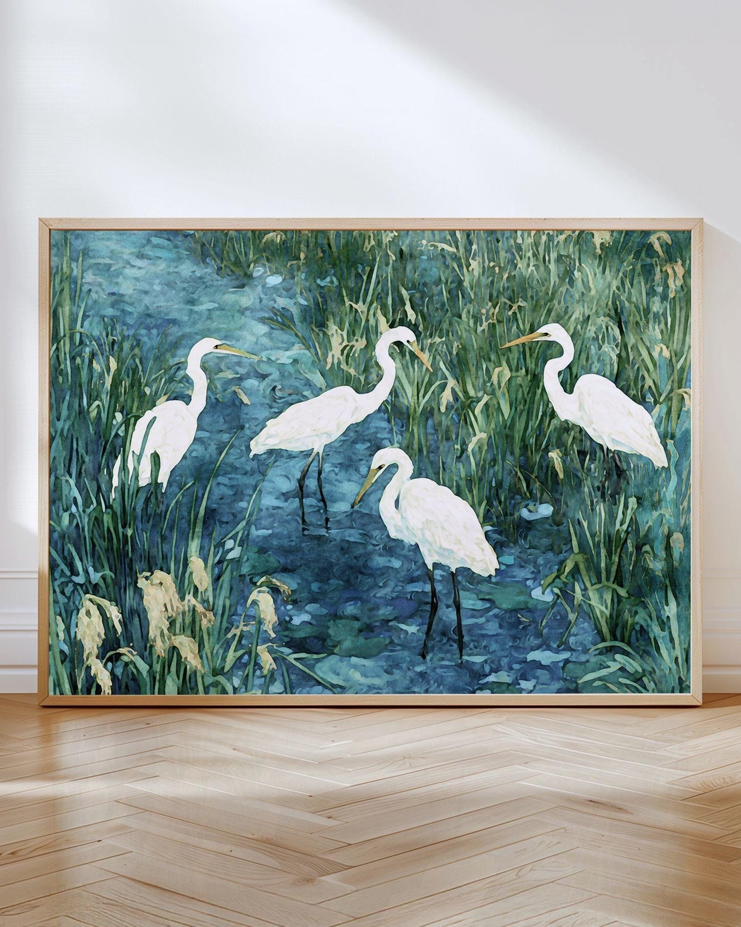 Framed painting of five white egrets standing in shallow blue water surrounded by tall green marsh grasses. The birds are depicted in soft watercolor-style brushstrokes, with elegant long necks and slender legs contrasting against the textured greenery and rippling water. The artwork rests against a white wall on a wooden herringbone floor, lit by natural sunlight.