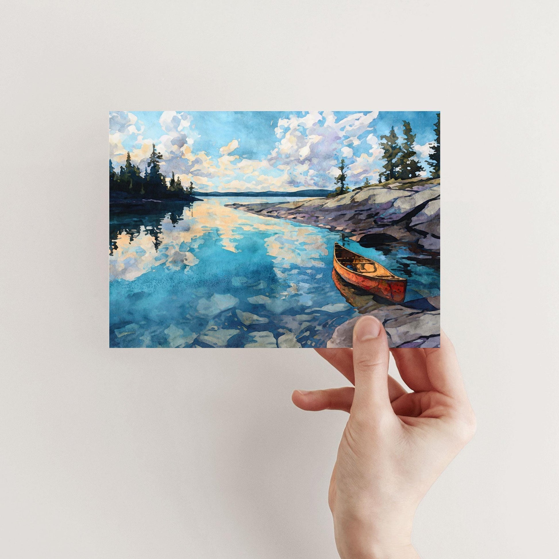 Hand holding a greeting card featuring a watercolor painting of a calm lake with a red canoe by rocky shore. The still blue water reflects golden clouds and pine trees along the horizon, evoking a peaceful wilderness scene.