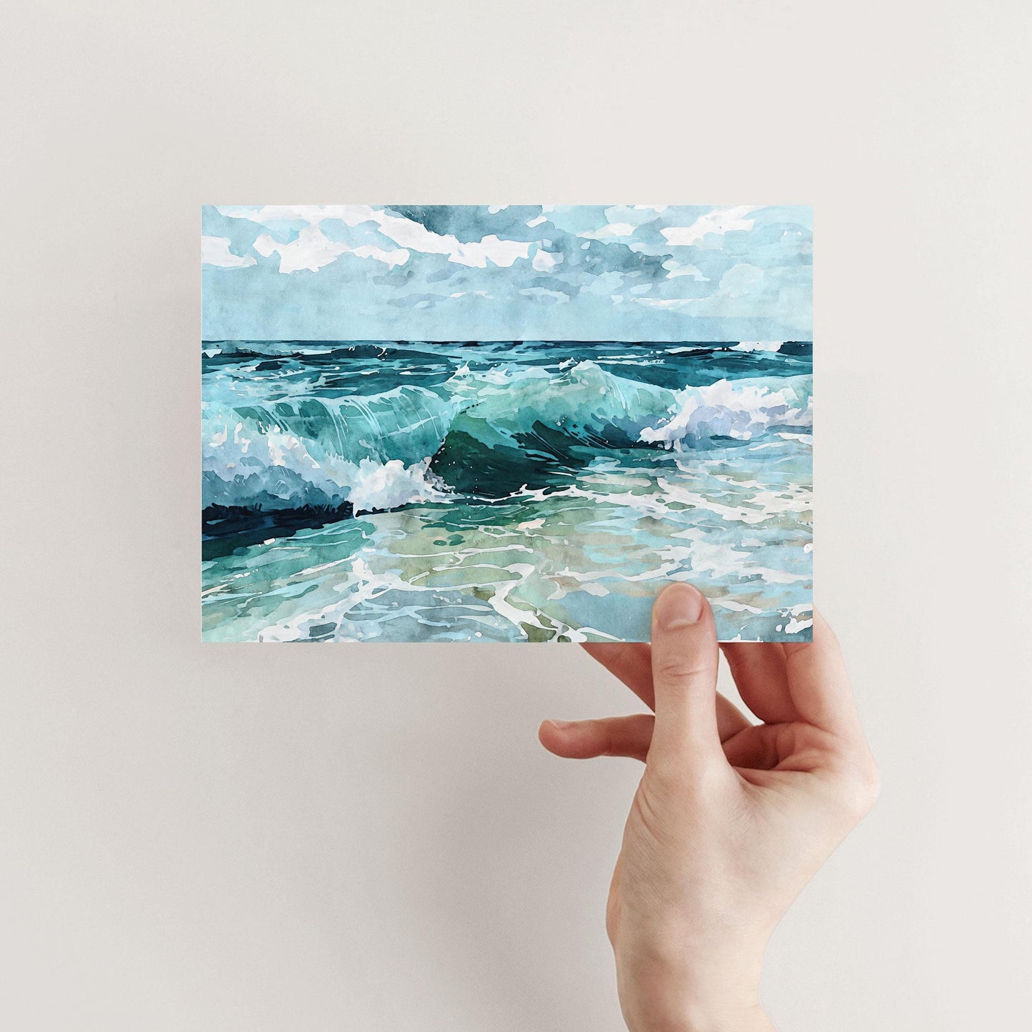 Hand holding a greeting card featuring a watercolor seascape with turquoise ocean waves crashing onto the shore beneath a partly cloudy blue sky.