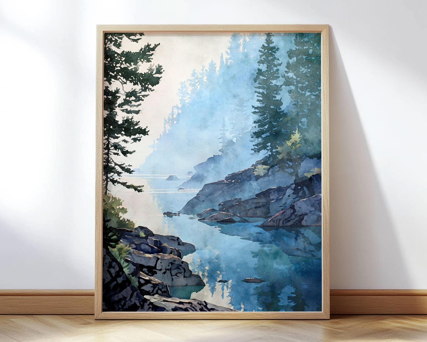 Framed watercolor-style landscape art print of a misty lakeshore with calm blue water reflecting evergreen trees and rocky shoreline. Soft morning fog drifts through tall pine trees along a quiet inlet, with layered blues and greens creating a serene, atmospheric forest scene. Vertical wall art shown in a light wood frame against a neutral wall.