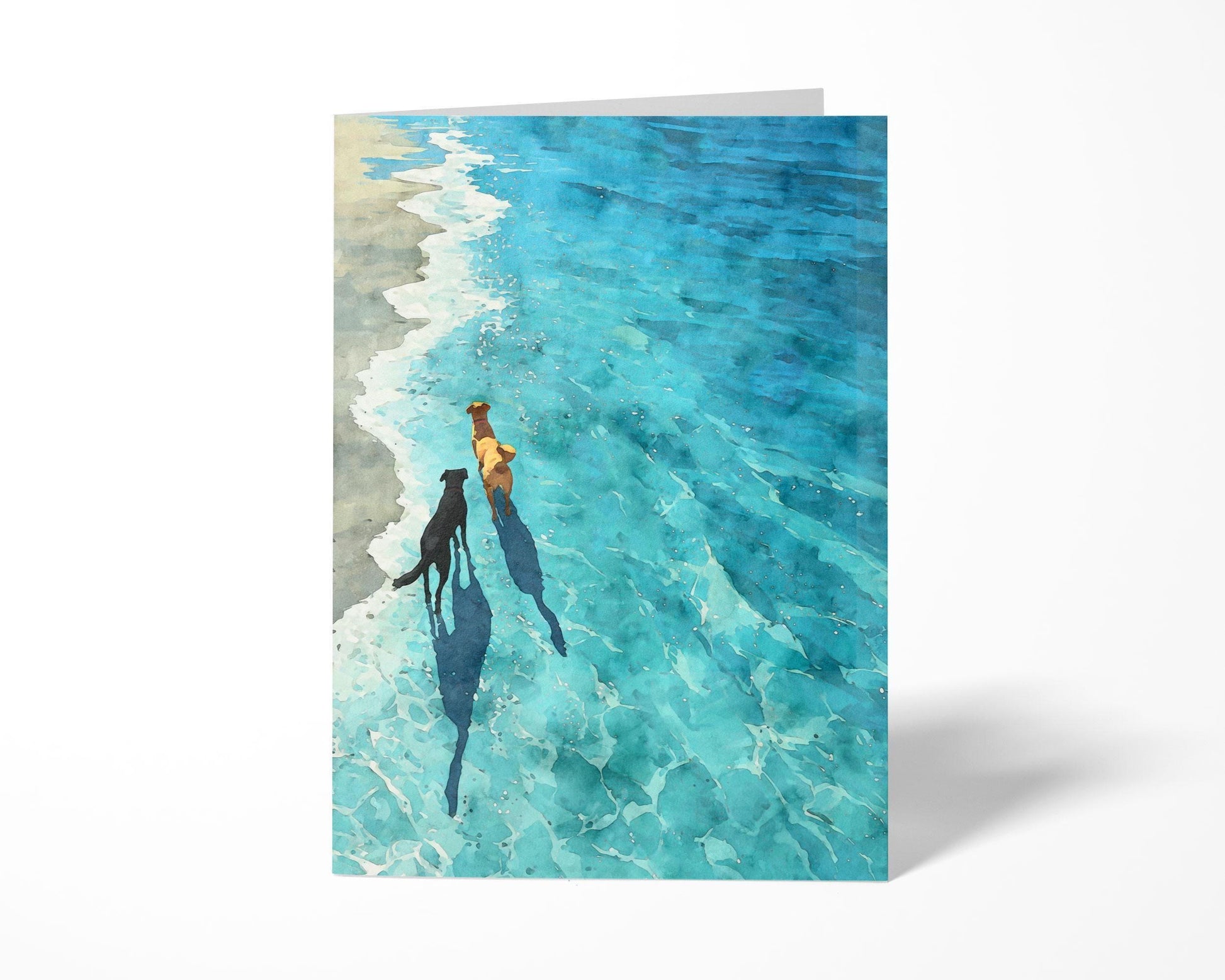 Greeting card standing upright on a white background, featuring a watercolor-style illustration of two dogs walking along the shoreline. The clear turquoise water and gentle waves create a peaceful beach scene, with long shadows cast by the dogs enhancing the sunny, tranquil mood.