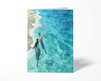 Greeting card standing upright on a white background, featuring a watercolor-style illustration of two dogs walking along the shoreline. The clear turquoise water and gentle waves create a peaceful beach scene, with long shadows cast by the dogs enhancing the sunny, tranquil mood.