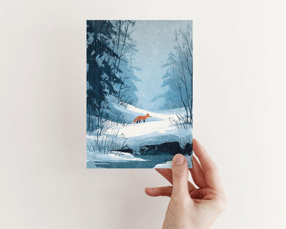 A hand holds a greeting card featuring a digital painting of a red fox walking through a snowy forest. The serene winter landscape is rendered in soft blue tones with snow-covered trees, bare branches, and gentle snowfall, creating a calm, atmospheric scene of quiet wilderness.