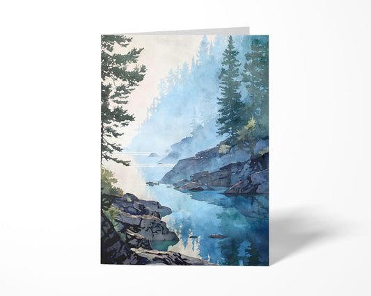 Vertical art greeting card featuring a watercolor-style misty lakeshore scene. Calm blue water reflects evergreen trees and rocky shoreline, softened by light morning fog. Painted in layered blue and green tones with visible watercolor texture, shown upright on a clean white background to highlight the artwork and card shape.