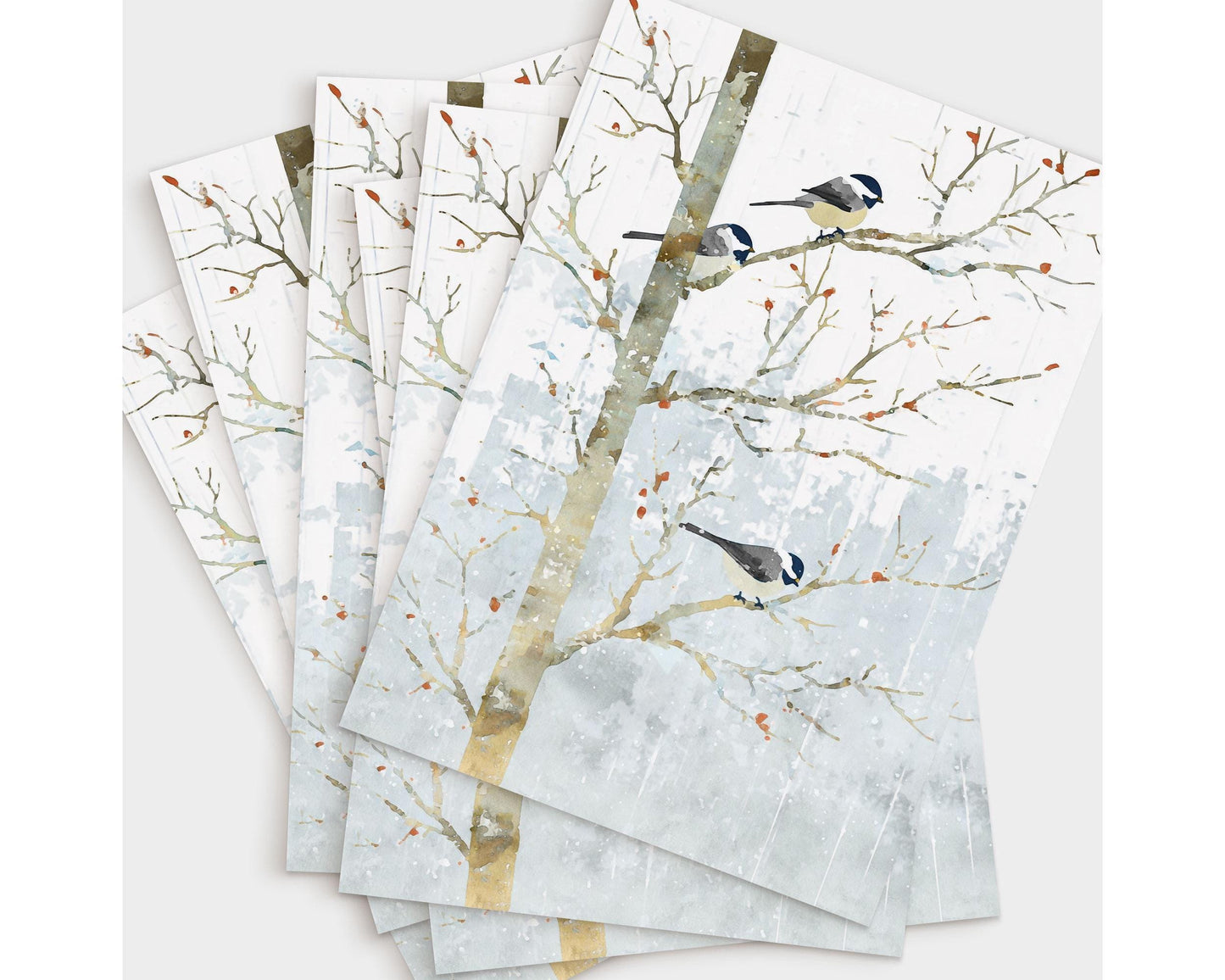 Set of greeting cards featuring a watercolor-style illustration of three chickadees perched on bare winter branches. The cards are fanned out on a light background, displaying soft gray-blue snowy tones and small orange berries for a peaceful winter look.