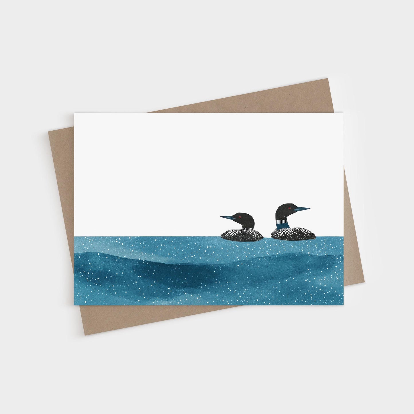 Minimalist greeting card with two loons illustrated in black and white, floating on a deep blue, star-speckled water pattern against a clean white background, paired with a kraft envelope.