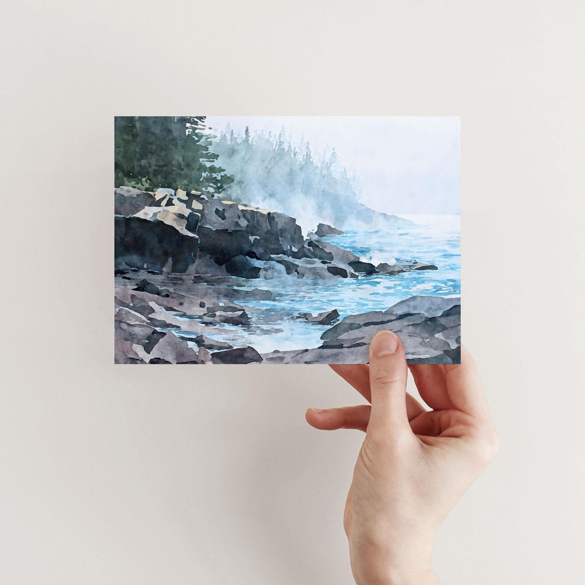 Horizontal greeting card shown held in a hand against a neutral background, featuring a watercolor-style illustration of a misty Lake Superior shoreline. Dark rocky cliffs and scattered stones line calm blue water, with soft waves and reflections in the foreground. Pale fog drifts across the scene, partially obscuring a distant evergreen forest, creating a serene, atmospheric coastal mood.